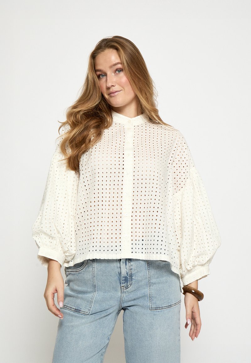 Blusa blanca con perforaciones, cuello alto, cierre de botones y mangas abullonadas. Combinada con jeans azul claro de talle alto.