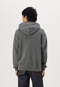 Grå huvtröja med avslappnad passform. Har mjukt tyg, ribbade muddar och en stor söm på baksidan för extra detalj. Bärs med mörka jeans.