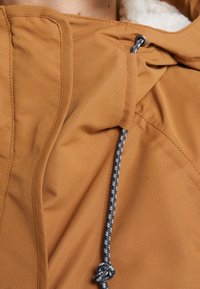 Brązowa kurtka z miękką, teksturowaną podszewką, z kapturem na sznurku i kontrastowym czarnym i białym sznurkiem. Widoczne szwy oraz kieszeń.