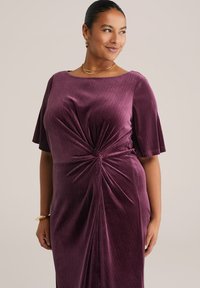 Szilva színű bársonydressz, testhezálló fazonnal, rövid, kibővülő ujjakkal és csavart csomóval a deréknál. Simás, puha tapintás.