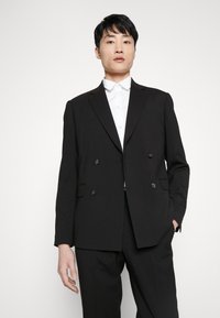 Blazer negro de doble botonadura con solapas de muesca, que presenta botones negros y bolsillos laterales. Llevado sobre una camisa blanca con cuello.