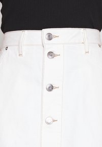 Gonna in denim bianco con cinque bottoni metallici argento sulla parte frontale, con dettagli di cucitura sottili e un tessuto texture.