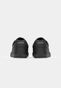 Črne superge z gladko mat površino, opremljene z zlato odtisnjeno oznako "LACOSTE" na peti in teksturirano gumijasto podplato.