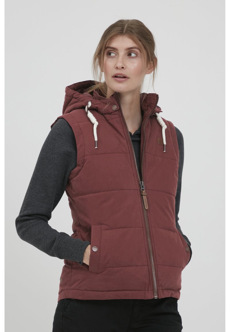 Gilet matelassé rouge avec une capuche, doté d'une fermeture éclair à l'avant, d'un cordon de serrage et de poches latérales. Porté par-dessus une chemise à manches longues grise.