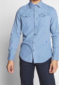 Ljusblå skjorta med lång ärm och nedknäppt krage, tvilling bröstfickor och subtil textur; har kontrasterande knappar och rundad nederkant.