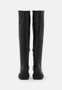 Botas negras de piel sintética hasta la rodilla con suela plana, cremalleras laterales y grandes anillas metálicas en la parte posterior para dar acento y un ajuste seguro.