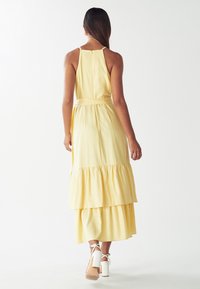 Geel tiered maxi-jurk met een halternek en een ritssluiting aan de achterkant. Glad stof met een cinched taille en een bijpassende strik. Witte sleehakken.