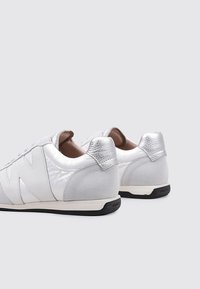 Zapatillas blancas con acentos reflectantes plateados, parte superior sintética suave, talón texturizado y suela de goma negra. Diseño simple, perfil bajo.