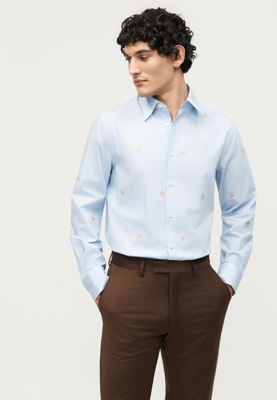 Άνδρας με γαλάζιο πουκάμισο με μικρά λουλουδάτα σχέδια και καφέ παντελόνι, με τα χέρια στις τσέπες, κοιτάζει αριστερά σε απλό φόντο.