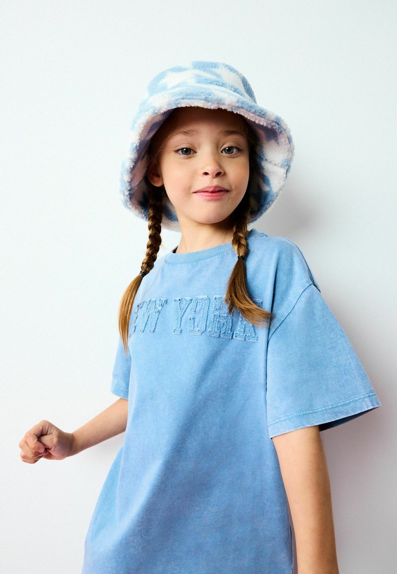 T-shirt en coton bleu clair avec "NEW YORK" brodé dans un fil assorti. Accompagné d'un chapeau seau en polaire avec un motif nuage bleu et blanc.