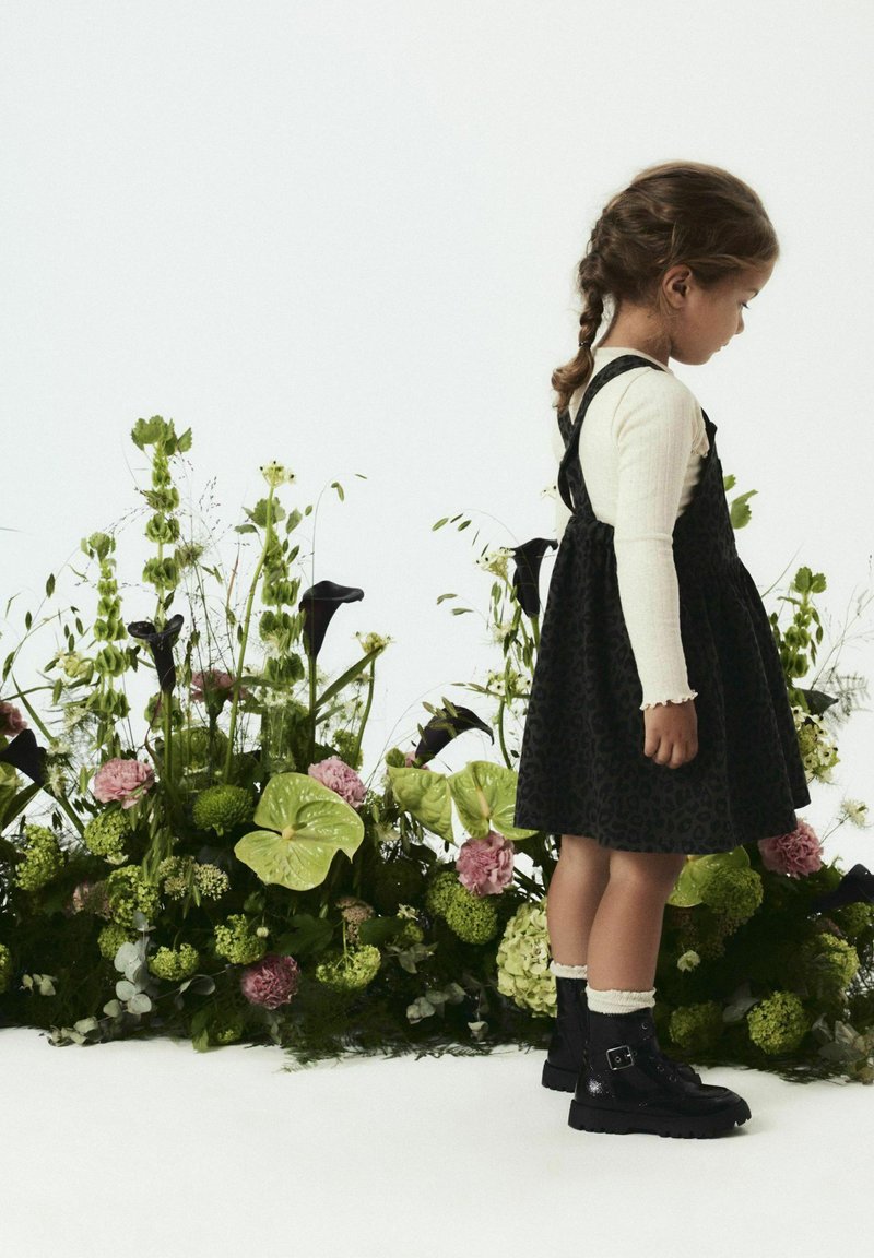 Schwarzes Leopardenmuster-Kleid mit einem ausgestellten Rock, weißem Langarmhemd und schwarzen Schnürstiefeln. Umgeben von einem bunten Blumenarrangement.