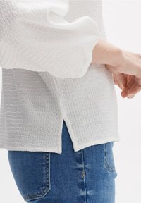 Haut blanc texturé avec de longues manches bouffantes et des fentes latérales, associé à un jean bleu, affichant un design décontracté et un tissu doux.