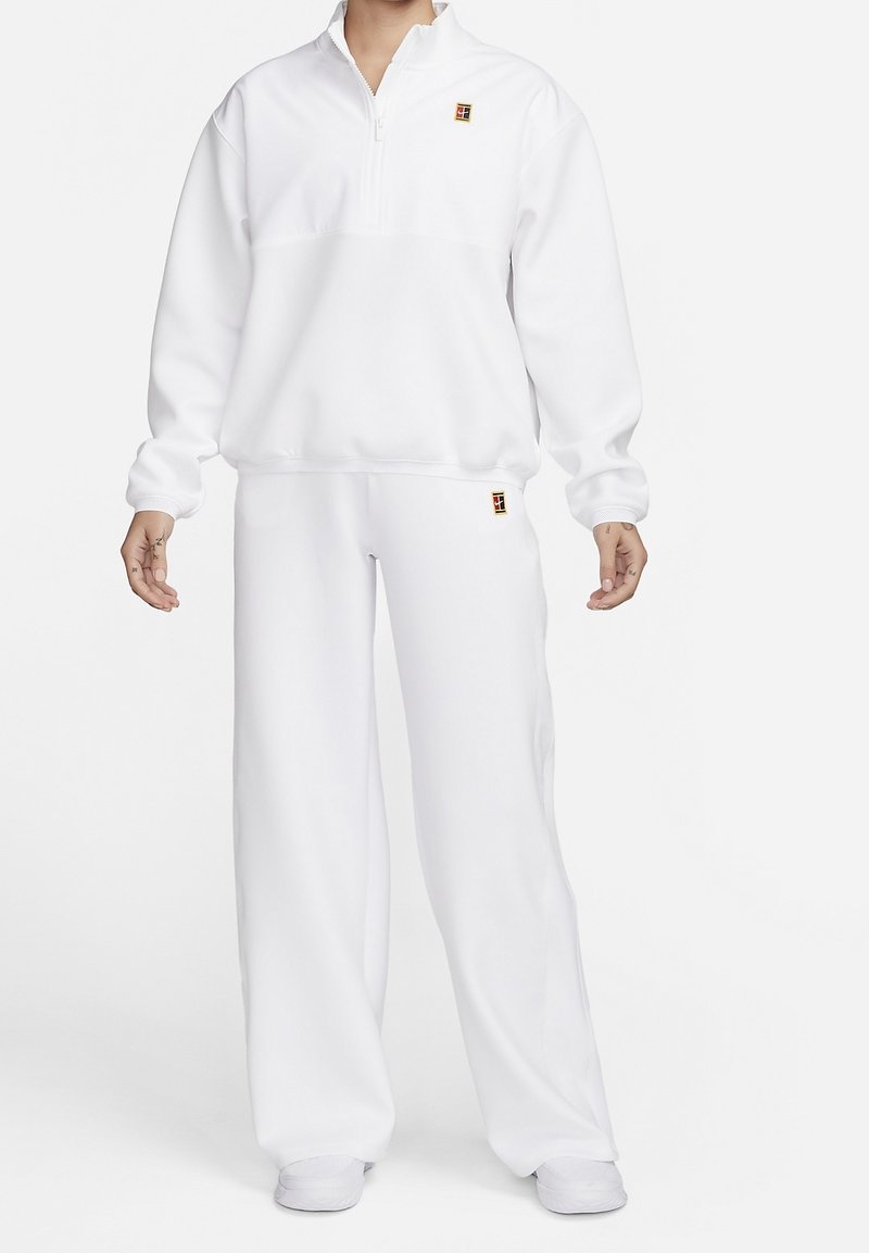 Valkoinen fleece-juoksuasu, jossa on neljännesvetoketjukaulus, pitkät hihat ja rento malli housuissa. Housuissa ja rinnassa on pieni värillinen logo.