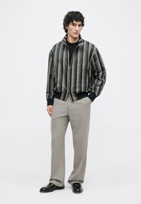 Mann mit lockigem Haar, der eine schwarz-grau gemusterte Reißverschlussjacke, hellgraue Hosen und schwarze Schuhe trägt, während er mit den Händen in den Taschen steht.