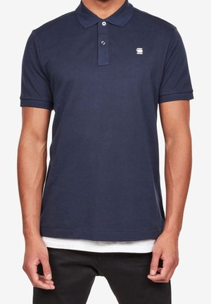 Homme portant un polo bleu marine à manches courtes avec un petit logo blanc sur le côté gauche de la poitrine et un pantalon noir.