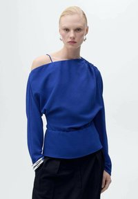 Top blu lungo con una spalla scoperta, dettaglio arricciato sulla spalla e fascia vita arricciata. Il tessuto appare liscio e testurizzato.