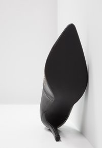 Chaussure à talon haut noire à bout pointu reposant sur le côté avec des clous dorés le long du bord supérieur sur fond blanc.