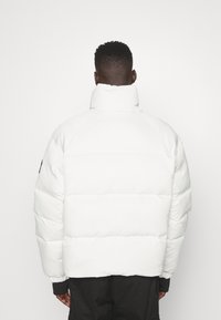 Uomo con capelli corti indossa un piumino bianco a collo alto e pantaloni neri, in piedi di spalle contro uno sfondo bianco uniforme.