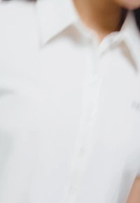 Valge nööpidega särk klassikalise kraega, millel on sile tekstuur, peidetud nupud ja diskreetne logo rinnal.