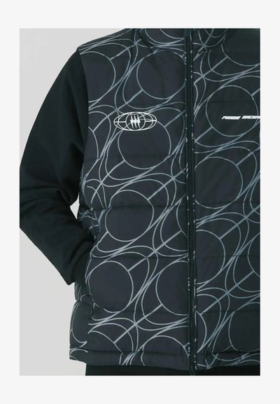 Gilet nero imbottito con motivi circolari argentati, chiusura con zip frontalmente e logo sul petto. Abbinato a una maglietta nera a maniche lunghe sotto.