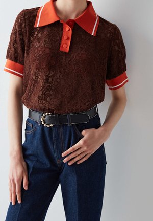 Personne portant un polo en dentelle marron avec col et poignets orange, rentré dans un jean bleu avec une ceinture noire à boucle décorative.
