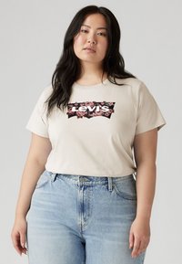 Beige t-skjorte med kort erm og en blomsterdekorert Levi's-logo på forsiden. Kombinert med lyseblå denimjeans. Glatt tekstur, avslappet passform.