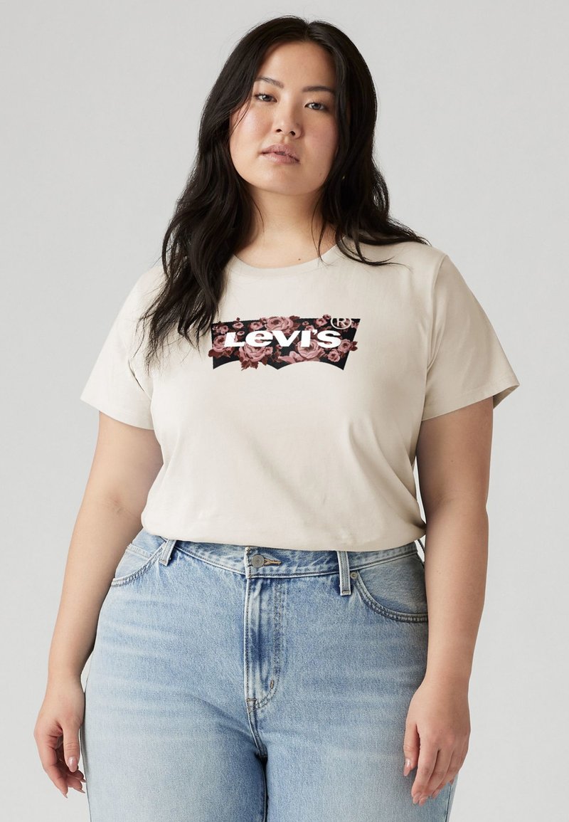 Beige t-skjorte med kort erm og en blomsterdekorert Levi's-logo på forsiden. Kombinert med lyseblå denimjeans. Glatt tekstur, avslappet passform.