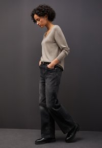 Donna con capelli ricci che indossa un maglione beige a scollatura a V, jeans neri a gamba larga e scarpe nere, in piedi su uno sfondo grigio scuro con le mani in tasca.