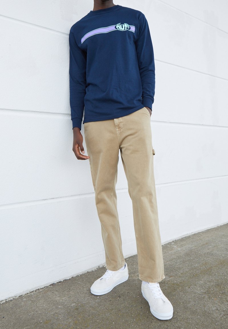 Chemise à manches longues bleu marine avec un motif graphique, associée à un pantalon beige et des baskets blanches. Tissu lisse et coupe décontractée.