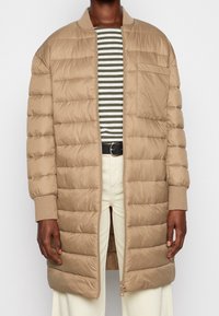 Béžová péřová bunda se zipem vpředu, žebrovanými manžetami a horizontálním prošíváním. Nasažená přes pruhované tričko a vysokým pasem krémových kalhot.