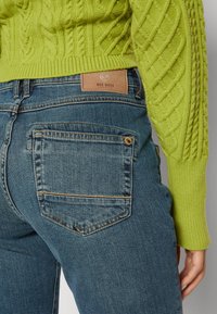 Jeans de mezclilla con un ajuste de tiro medio, en color azul. Presentan una etiqueta de cuero color marrón, bolsillos traseros y un suéter de punto en color verde lima.