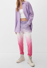 Fioletowa koszula zapinana na guziki, biała crop top, gradientowe dżinsy przechodzące od jasnego fioletu do różu oraz beżowe sneakersy za kostkę.