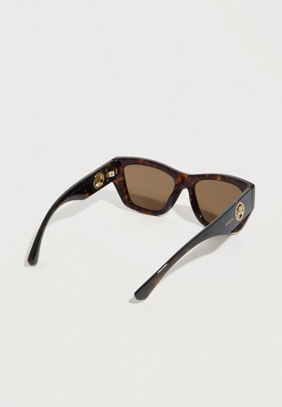 Καφέ ορθογώνια γυαλιά ηλίου με σχέδιο χελωνονόπαρου, σκούρους φακούς και χρυστό στρογγυλό λογότυπο μάρκας στους βραχίονες, τοποθετημένα πάνω σε λευκή επιφάνεια.