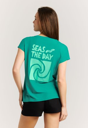 Femme en t-shirt turquoise avec l'inscription « SEAS THE DAY » et un motif de vague dans le dos, portant un short noir, debout de dos à la caméra.