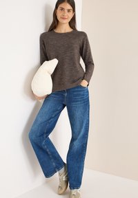 Maglione lungo a maniche e melange marrone, jeans blu larghi e sneakers grigi. Tiene un cuscino crema trapuntato, in piedi contro uno sfondo neutro.