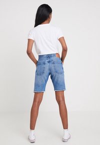 Blå denimshorts med fransad nederkant, fem fickor och en röd etikett baktill. Matchas med en enkel vit t-shirt och vita sneakers.