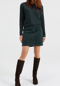 Maglione verde a coste abbinato a una minigonna in suede verde scuro con bottoni frontali. Stivali in suede marrone al ginocchio completano l'outfit.