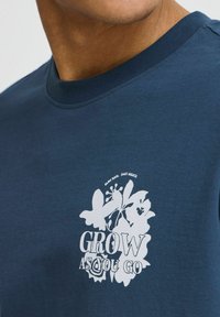 Mörkblå t-shirt med kort ärm i bomull. Har en vit blommig grafik med texten "GROW AS YOU GO" i fet stil.