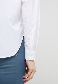 Camicia bianca in cotone a maniche lunghe con polsini abbottonati, caratterizzata da un orlo curvo e cuciture visibili, abbinata a pantaloni in denim blu.