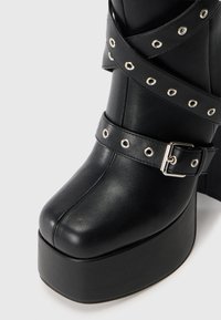 Zwarte plateauboots met een strakke leertextuur, voorzien van gekruiste riemen met zilveren oogjes en een gesp-accent. Vierkante neusdesign.