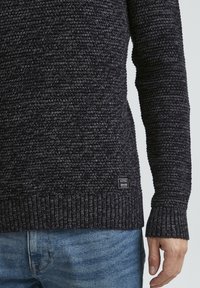 Sötét szürke kötött pulóver, bordázott mandzsettákkal és aljjal, textúrált mintával és kis márkajelzéssel az alsó bal oldalon.