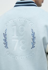 Ανοιχτό μπλε μπουφάν με ανάγλυφο κείμενο "Carlo Colucci 1978" και μπλε κεντημένα κλαδιά δάφνης στην πλάτη, φορεμένο από άτομο που κοιτάει πλάγια.