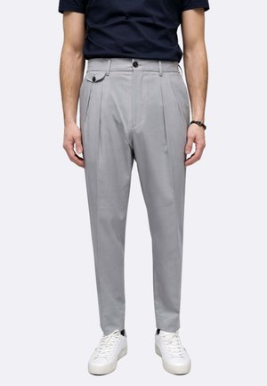 Uomo che indossa pantaloni plissettati grigio chiaro, sneakers bianche, cintura nera e una camicia a maniche corte blu scuro, in piedi davanti a uno sfondo uniforme.