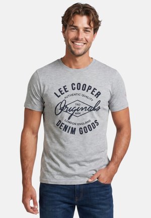 Homme souriant portant un t-shirt gris clair « Lee Cooper Originals Denim Goods » et un jean bleu foncé, debout avec une main dans la poche.