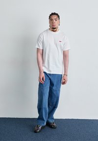 T-shirt de algodão branca com logótipo vermelho no peito, acompanhada de jeans de denim azul médio e soltos, e ténis pretos com detalhes verdes.