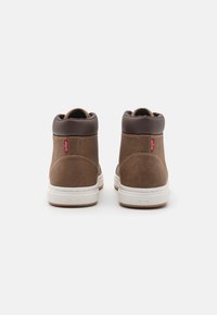 Zapatillas marrones de media caña con material texturizado, acentos en marrón oscuro, suela de goma blanca y una etiqueta roja de Levi's en la parte trasera.