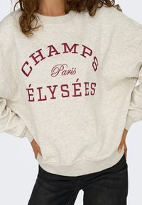 Ljust grå sweatshirt med maroon text som säger "CHAMPS PARIS ÉLYSÉES", med avslappnad passform och ribbade muddar och fåll. Kombineras med mörka jeans.