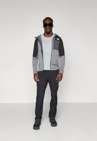 Chaqueta con cremallera en gris claro y negro con capucha, combinada con una camiseta azul claro y pantalones negros; el conjunto se completa con zapatillas negras y gafas de sol.
