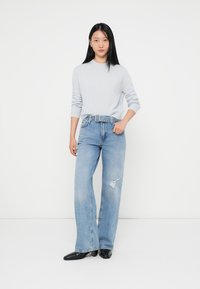 Lichtblauwe lange mouwen trui gecombineerd met hoge taille, wijde spijkerbroek met slijtage. Model draagt zwarte pumps met spitse neus en staat tegen een witte achtergrond.