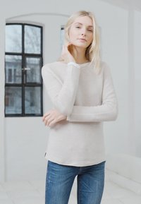 Haut beige clair à manches longues avec une texture côtelée, col rond et bordure blanche contrastante. Associe avec un jean bleu. Splendide éclairage intérieur.